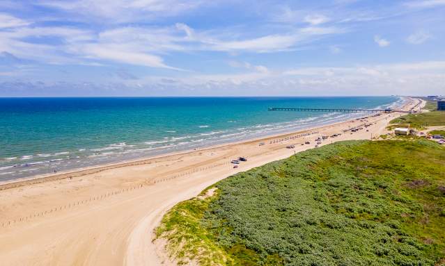 Drone image of a beach that shows the water, beach, and sand dunes from very high. Down the beach, a few cars and a pier are visible.