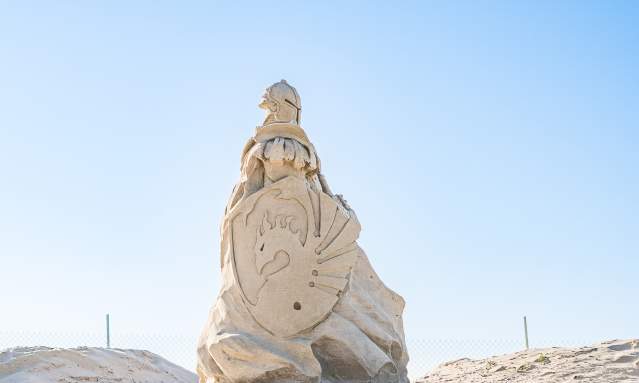 Sand sculpture on the beach of a guardian soldier facing the left. The soldier carries a shield emblazoned with a dragon.