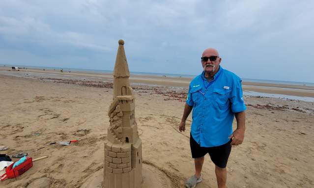 A sculptor in a blue fishing shirt stands on the beach next to a sand sculpture.