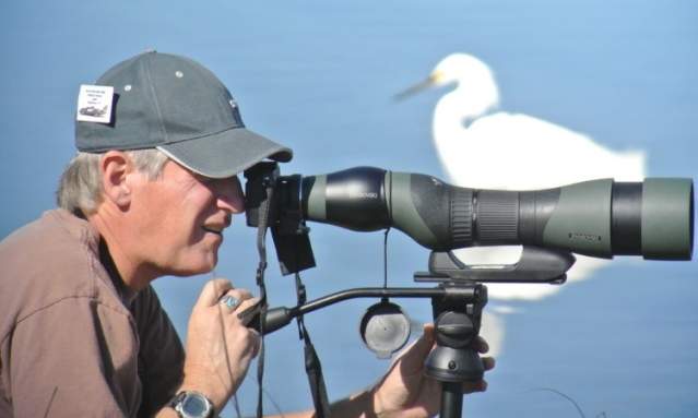 A man in a cap looks through a telescope with a large white bird in the background.