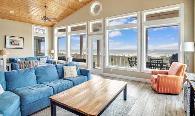 Photo of a living room interior with ceiling to floor glass windows looking out to the beach. An orange chair, blue couches, lamps, and coffee tables are visible.