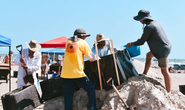 A group of four men crowd around forms on a pile of sand with buckets and sculpting tools