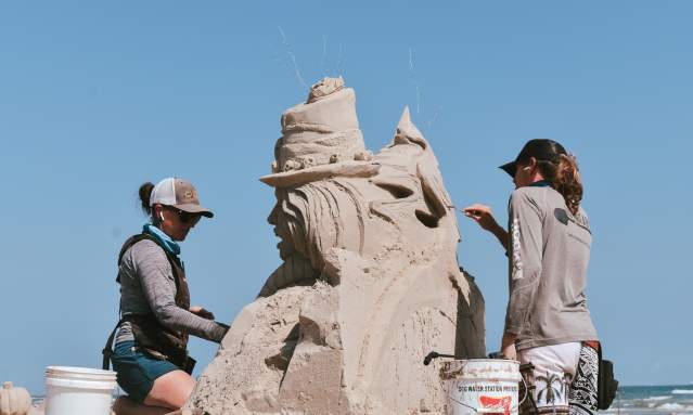 Two people, a man and a woman, stand on either side of a sand sculpture and carve details.