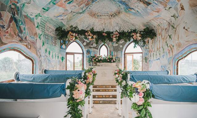 The interior of a chapel painted with murals is decorated with pink, white, and green florals, and white pews are draped in light blue fabric