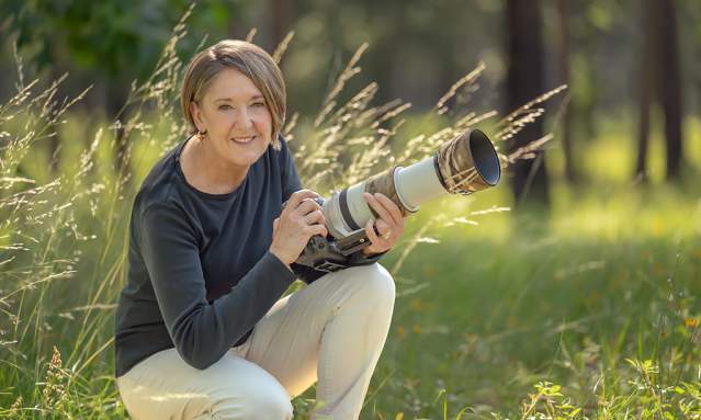 A woman smiles while kneeling in a field with a large camera