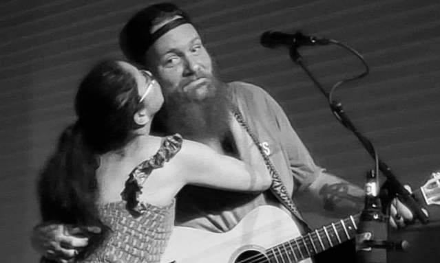 Black and white photo of a bearded man holding a guitar being kissed on the cheek by a woman in a long dress.