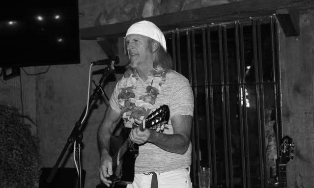 Black and white photo of a man wearing a lei strumming a guitar and singing into a microphone.