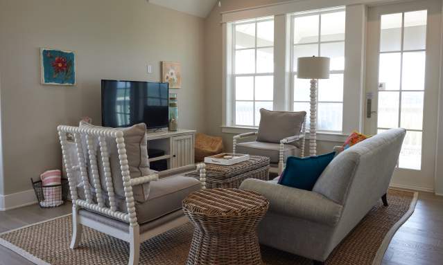 A living room with coastal decor in shades of beige. A tan couch faces a television and two chairs face each other perpendicular to the couch.