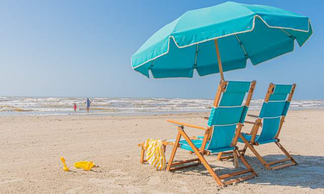 Two blue beach chairs with an unbrella between them. There is a yellow blanket over one chair and yellow beach toys near by. In the background thier are two people playing in the ocean.