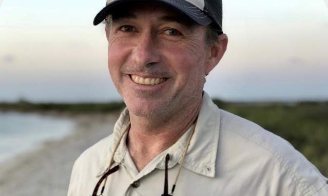 A man in a tan shirt and a hat smiles at the camera. There is a beach in the background.