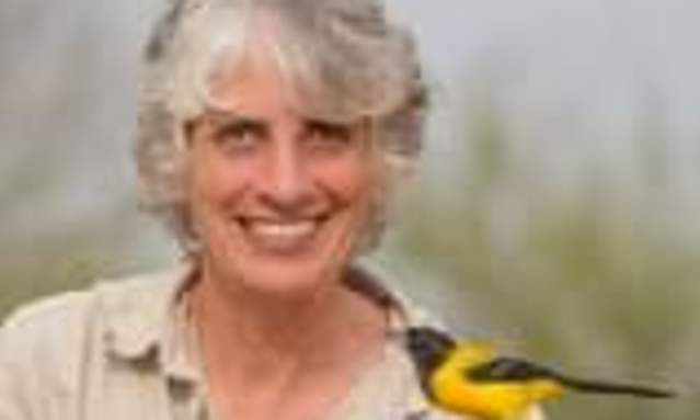 A middle-aged woman with short in a tan shirt holds a yellow and black bird on her hand