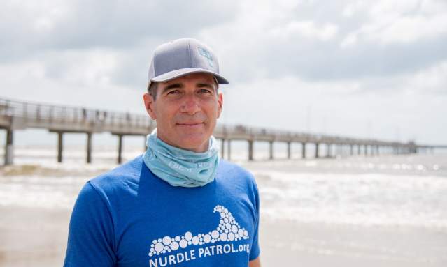 A man in a blue shirt reading "Nurdle Patrol" and a hat stands on the beach and smiles at the camera.