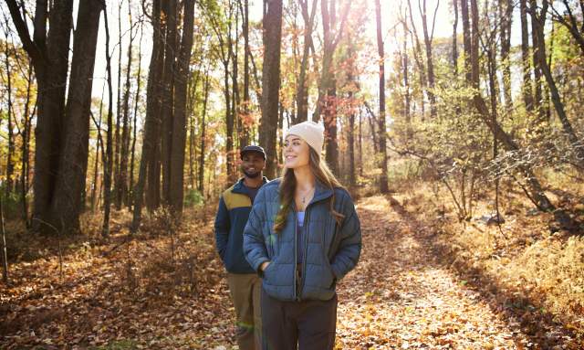 A couple walks on a trail in a forest with fallen brown and orange leaves wearing warm jackets and hats on a fall day.