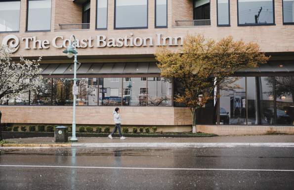 A person walks past The Coast Bastion Inn on a rainy day, with wet pavement and trees lining the street.