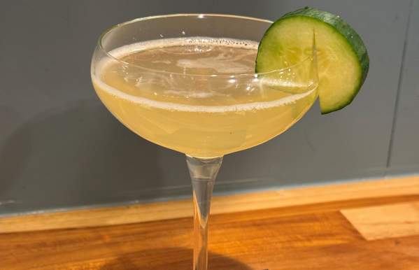 A coupe glass filled with a pale yellow cocktail, garnished with a fresh cucumber slice, placed on a black napkin atop a wooden bar counter with a muted blue-gray wall in the background.