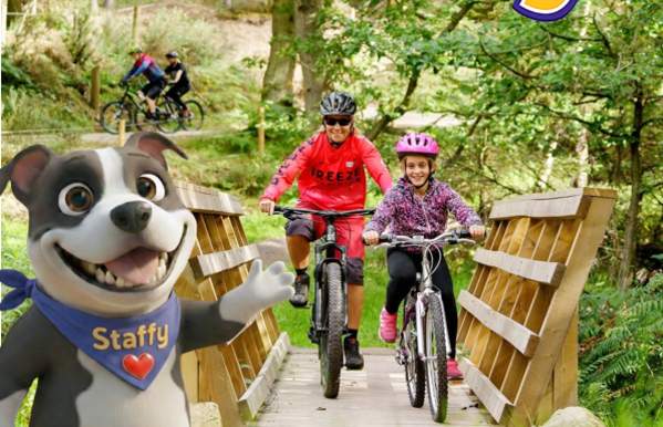 Infographic with Staffy visitor mascot in the foreground, adult and child riding bikes across a wooden bridge at Cannock Chase and Scholl's Out for Staffy logo.