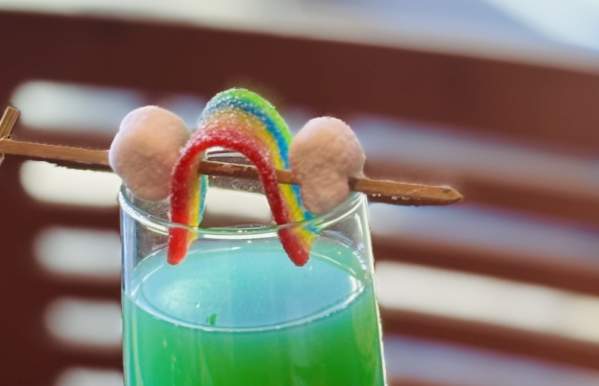 A festive green cocktail served in a champagne flute, garnished with a rainbow candy and marshmallows on a skewer. The drink sits on a wooden table surrounded by gold-wrapped chocolates, creating a fun and whimsical presentation.