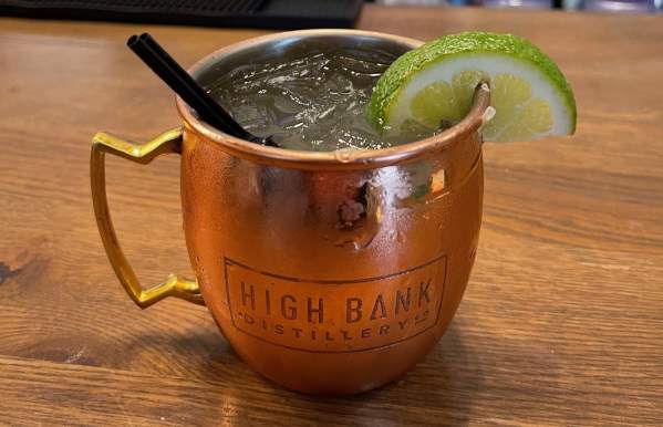 "A copper mug with 'High Bank Distillery Co.' embossed on the side, filled with an ice-cold cocktail, garnished with a fresh lime wedge and a black straw, sitting on a wooden bar counter with a blurred background of a bar setup