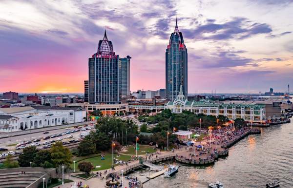 A vibrant sunset over Downtown Mobile skyline, featuring tall buildings, a bustling waterfront, and a lively crowd below.