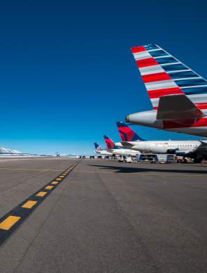 Airplanes lined up on a runway with snow-capped mountains in the background under a clear blue sky. The scene captures a vibrant travel hub in Jackson Wyoming.