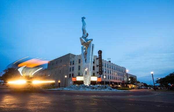 steel sculpture "muskegon rising" sits in roundabout in front of historic Frauenthal theater at sunset. blurred car headlights illuminate the left side of the photo