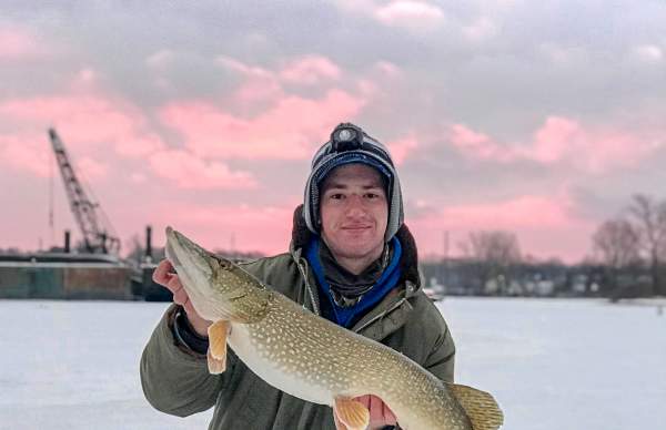 A person in winter attire holding a large Northern Pike while kneeling on a frozen Muskegon Lake.
