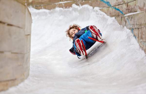 young boy crests curve of snow covered luge track on luge sled