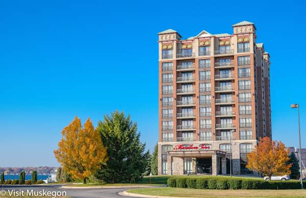 Hotel exterior on Muskegon Lake. The sky is blue and the trees have autumn color.