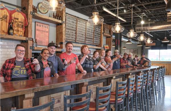 A group of people at a brewery bar raise small beer glasses in a toast. They stand by a long wooden counter with menus on the wall, looking cheerful.
