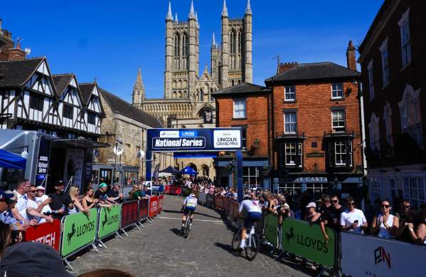 Two cyclists compete in the Men's Tour of Britain race in Lincoln, with spectators watching from behind crowd barriers on either side of the route.