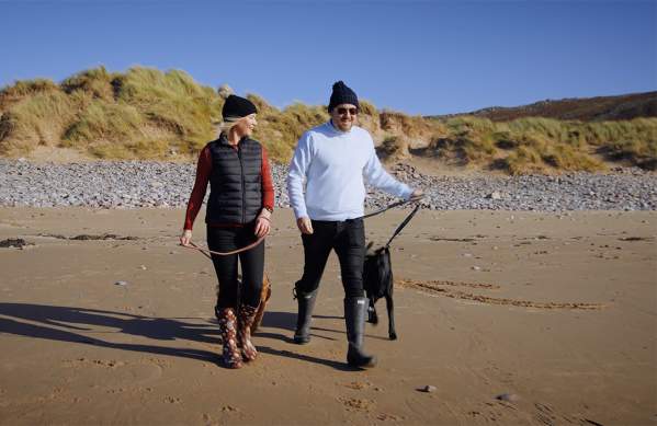 A man and woman wearing warm clothes, each walking a dog on a beach.