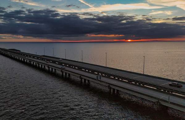 Aerial view of the Lake Pontchartrain Causeway Bridge at dusk, stretching southwest across Lake Pontchartrain from the Northshore toward Metairie. Vehicles travel along the twin spans beneath dramatic dark clouds, while a sliver of orange sunset glows along the horizon and reflects faintly on the calm water.