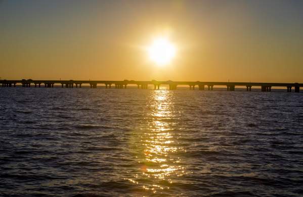 The Lake Pontchartrain Causeway Bridge stretches across Lake Pontchartrain at sunset, silhouetted against a golden sky. The low sun hangs above the bridge, casting a bright reflection across the rippling water as vehicles travel along the span between the Northshore and the Southshore.