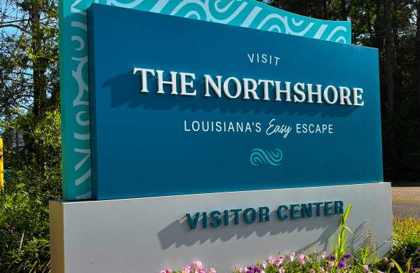 Visit The Northshore Visitor Center sign beside Highway 59 in Mandeville, Louisiana, surrounded by blooming flowers and landscaped greenery.
