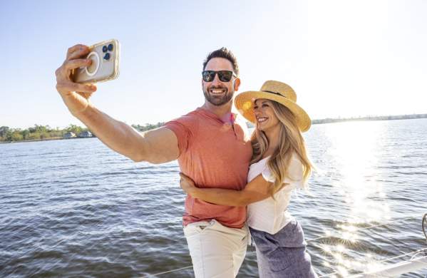 A man and a woman stand on a sailboat near sunset. The man is taking a photo of himself and the woman.