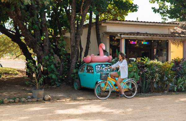 Como moverse en Panamá, mujer recorriendo Playa Venao en bicicleta pasando junto a un coche azul vintage con un flotador de flamenco rosa