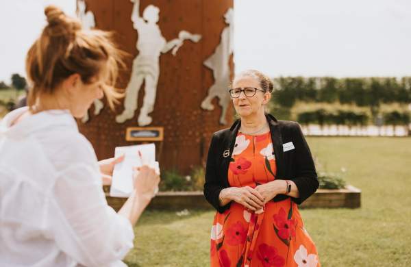 Woman in red dress looking at woman in white shirt in front of a steel sculpture outdoors.