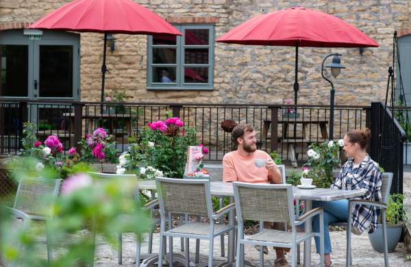 A man and a woman sat on a outdoor dining table with hot drinks in courtyard with listed barn building in background
