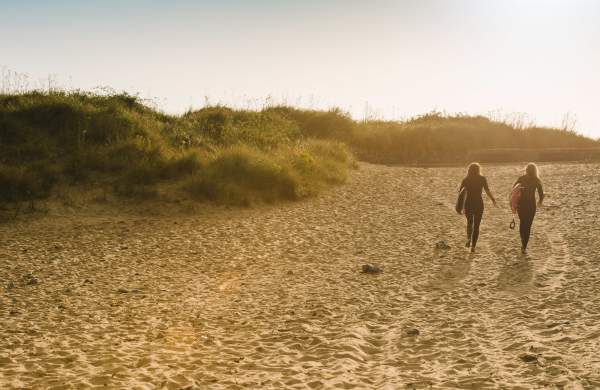 Two ladies walking across the beach with surfboards