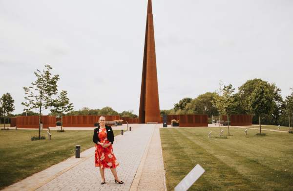 a woman stood on a path surrounded by fields and trees with a spire monument in the background