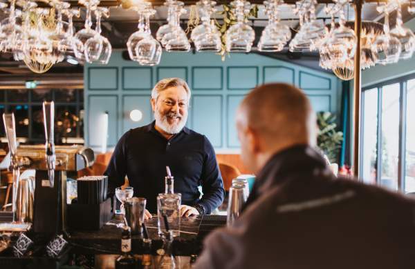 a man looking at bar staff over a bar with glasses hanging from a rack
