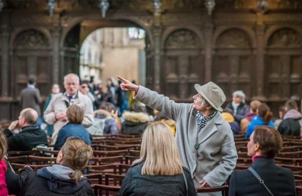 White haired white woman in a grey wool coat & grey felt hat, standing profile pointing to the left in Kings College Cambridge Chapelpointing