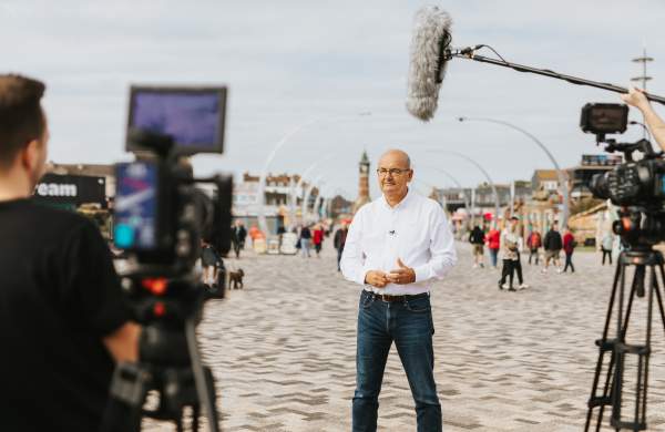 A man stood on skegness promenade with a clock tower in the background and a camera crew out of focus in the foreground