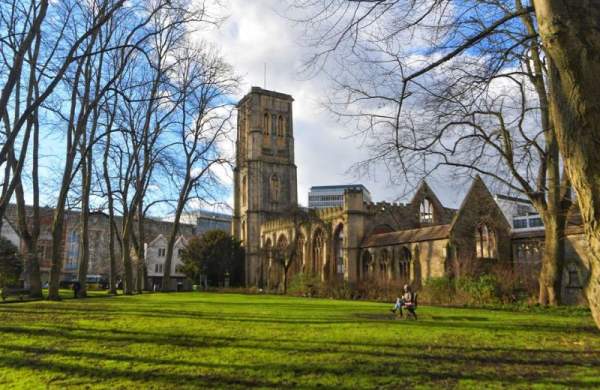 An image showing Temple Church Gardens Quiet Area and the green grass of the gardens in front. There are trees around the edge of the grass and a person sitting on a bench.