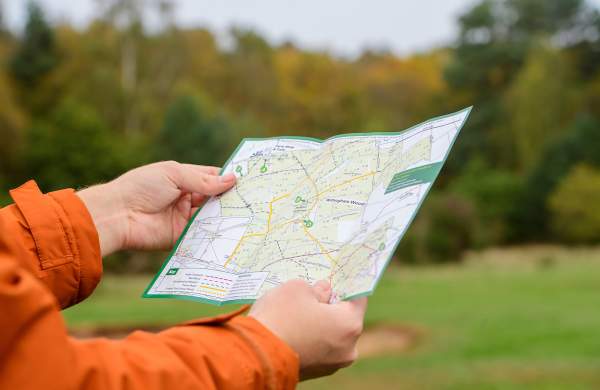 a close up of a man holding a walking guide map with a bright orange coat on and autumnal trees in background