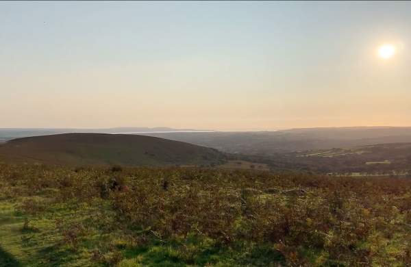 A rural Welsh landscape with the sea in the distance and a low sun in the sky.