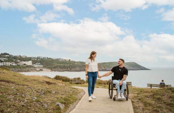 A man in a wheelchair holding hands with a standing woman on a section of the coast path with the sea in the background.