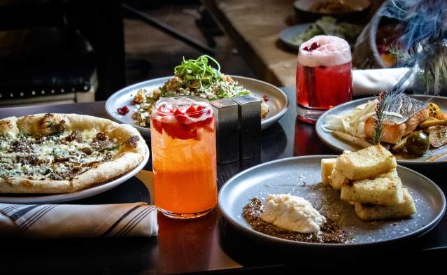An array of food and drink on a table top that includes pizza and some colorful cocktails.