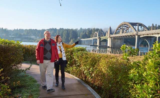 A couple walks near the Siuslaw River in Historic Old Town Florence with the beautiful highway 101 bridge in the background.