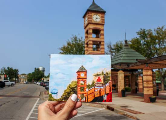 A handpainted postcard showing the downtown Overland Park clocktower is held up in front of the physical clocktower.